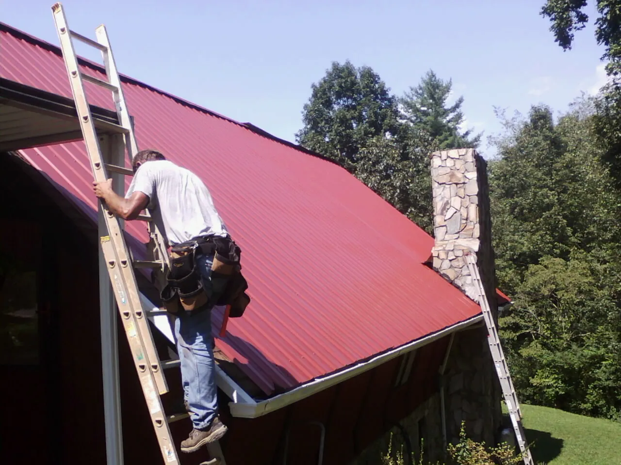 Roofer on a ladder beside a red metal roof and stone chimney — Soffit & Fascia Repair by Roofing Brothers in Danube